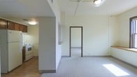 This single-floor space features a straightforward, functional layout with a clean, neutral aesthetic throughout. The main living area is finished with light grey carpet, providing a soft underfoot feel, while the adjacent kitchen transitions to hard flooring, offering a practical distinction between zones. The kitchen area, partially visible behind a partial dividing wall, includes a white range and cabinetry, creating a compact yet efficient cooking space. A built-in shelf on the dividing wall adds a small but useful architectural detail between the two areas. The living space is illuminated by ceiling-mounted light fixtures, ensuring consistent brightness throughout. A doorway with a dark-framed surround leads to an additional room, hinting at further functional space beyond. The walls are finished in a soft off-white tone, contributing to a bright and airy atmosphere. The ceiling features a ceiling fan, adding to the ventilation options within the space. Overall, the layout is efficient and practical, with clear zoning between living and kitchen areas, making it a versatile single-floor space suited to a variety of uses.