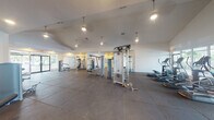 This single-floor fitness center offers a comprehensive workout environment thoughtfully organized across several distinct zones. The main exercise floor is expansive, featuring soaring vaulted ceilings with pendant lighting and large-format dark tile flooring that lends a clean, professional feel. This central area is outfitted with a variety of cable and strength training machines, providing ample space between equipment for comfortable movement.

A dedicated cardio zone hosts a row of treadmills, ellipticals, and a stationary bike, positioned alongside wide windows that frame lush outdoor greenery and what appears to be a pool area, keeping the space bright and energizing. A separate free weights area accommodates multiple dumbbell racks and an adjustable bench, bathed in natural light from sliding windows.

Adjacent to the main floor is a versatile aerobics and yoga studio with warm hardwood flooring, recessed lighting, and a clean, minimalist design. Glass doors connect this room to the rest of the facility, maintaining an open and airy atmosphere ideal for group classes or individual stretching sessions.

Throughout the facility, white walls and abundant natural light from large windows and glass doors create a bright, welcoming ambiance, making this fitness center a well-rounded destination for all types of training.