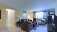 This single-floor space features a living room designed for comfort and functionality. The room is finished with light-colored carpeting that extends throughout, complementing the warm, neutral wall tones that create an inviting atmosphere. A large window dressed with dark curtain panels allows abundant natural light to fill the space, brightening the room while offering the option for privacy. Below the window sits a wall-mounted air conditioning unit, providing climate control convenience.

The living area is furnished with a plush, dark brown sofa accented with decorative pillows in muted tones, paired with a matching oversized chair that adds to the seating capacity. A low-profile rectangular coffee table anchors the seating arrangement atop a dark area rug, adding definition to the space. A cushioned accent chair with an ottoman provides an additional relaxation spot. Ambient lighting is offered through table lamps on side tables, contributing to a warm, layered lighting scheme. A framed wall piece adds a decorative focal point. An open doorway visible to one side suggests a connected layout, enhancing the overall flow of the space.