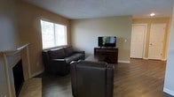 This single-floor space offers a welcoming and functional layout designed for comfortable everyday living. The living room serves as the heart of the space, featuring warm beige walls and durable hardwood-style flooring that runs throughout, lending a clean and cohesive look. A large window fitted with horizontal blinds allows generous natural light to fill the room, creating a bright and airy atmosphere. The living area is furnished with dark leather seating — including a sofa and matching chairs — arranged around a central coffee table, making it an ideal spot for relaxing or entertaining. A wooden media console with a flat-screen television anchors one wall, completing the entertainment setup. The textured ceiling adds a subtle decorative element that complements the overall neutral color palette. A doorway visible toward the back of the space hints at additional rooms, suggesting a practical and connected floor plan. Overall, the space presents a straightforward and comfortable environment with warm finishes, functional furnishings, and a layout that prioritizes ease of movement and livability.