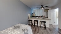 Located at 17839 Plummer St in the Northridge neighborhood of Los Angeles, this single-floor studio offers a thoughtfully designed open-concept layout that maximizes functionality within a compact footprint. The main living area is finished with wood-look plank flooring throughout, providing a cohesive and modern aesthetic that carries seamlessly from room to room.

The living and sleeping area is anchored by a ceiling fan with an integrated light fixture, providing both airflow and ambient illumination. Cool gray-toned walls lend a calm, contemporary feel to the space. A granite-topped breakfast bar serves as the central dividing element between the sleeping area and the kitchen, offering casual seating with three bar stools and built-in shelving beneath for additional storage.

The kitchen is equipped with a sink, countertop workspace, and small appliances, complemented by a wall-mounted air conditioning unit and vertical blinds over a window that allows natural light to filter in.

A separate area features elegant sliding frosted-glass doors with dark wood frames, concealing a built-in shelving and storage closet system. The bathroom is cleanly finished, maintaining the same wood-look flooring and light blue-gray wall tones found throughout the rest of the unit.