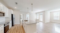 This thoughtfully designed single-floor residence offers a seamless blend of modern aesthetics and functional living. The open-concept kitchen and living area serves as the heart of the home, anchored by a large central island topped with granite countertops and fitted with an undermount sink. Pendant lighting with brass-toned fixtures illuminates the island, while crisp white shaker-style cabinetry, a subway tile backsplash, and stainless steel appliances — including a refrigerator with ice maker, electric range, and built-in microwave — complete the kitchen. Light hardwood flooring flows throughout the main living areas, reflecting natural light that pours in through multiple windows and a glass-paneled door in the living room. The bedroom features plush carpeting, dual windows with outdoor views, a ceiling fan, and easy access to a dedicated walk-in closet equipped with multi-tier wire shelving and a natural light window. The bathroom is finished with granite countertops, white cabinetry, and a glass-enclosed shower, maintaining the clean and cohesive aesthetic found throughout. Neutral warm-toned walls and recessed lighting across the space contribute to a bright, welcoming ambiance.