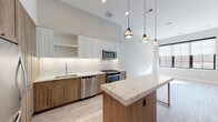 This thoughtfully designed single-floor residence showcases a seamless blend of modern finishes and warm, natural materials. The open-concept kitchen serves as the heart of the home, featuring a striking two-tone cabinetry scheme with crisp white upper cabinets and rich natural wood lower cabinets. A central island topped with veined quartz countertops provides additional workspace and casual dining, illuminated by a trio of industrial-style pendant lights overhead. The kitchen is equipped with a full suite of stainless steel appliances, including a gas range, built-in microwave, and dishwasher, all set against a classic white subway tile backsplash. Light-toned hardwood flooring runs throughout the living areas, enhancing the airy, open feel of the space. A standout detail is the rustic barn-style sliding door, which adds a charming farmhouse accent to an otherwise contemporary interior. The bathroom is well-appointed with a backlit vanity mirror, dark granite countertops, white cabinetry, and a full soaking tub with wood-look tile surround. Generous ceiling heights and large windows allow natural light to flow freely, creating a bright and welcoming ambiance throughout.