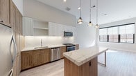 This thoughtfully designed single-floor residence showcases a seamless blend of modern finishes and warm, natural materials. The open-concept kitchen serves as the heart of the home, featuring a striking two-tone cabinetry scheme with crisp white upper cabinets and rich natural wood lower cabinets. A central island topped with veined quartz countertops provides additional workspace and casual dining, illuminated by a trio of industrial-style pendant lights overhead. The kitchen is equipped with a full suite of stainless steel appliances, including a gas range, built-in microwave, and dishwasher, all set against a classic white subway tile backsplash. Light-toned hardwood flooring runs throughout the living areas, enhancing the airy, open feel of the space. A standout detail is the rustic barn-style sliding door, which adds a charming farmhouse accent to an otherwise contemporary interior. The bathroom is well-appointed with a backlit vanity mirror, dark granite countertops, white cabinetry, and a full soaking tub with wood-look tile surround. Generous ceiling heights and large windows allow natural light to flow freely, creating a bright and welcoming ambiance throughout.