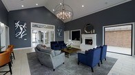 This single-floor space makes a striking impression through its thoughtful design and refined aesthetic. The centerpiece of the main lounge area is a dramatic vaulted ceiling from which an oversized orb chandelier descends, casting warm, ambient light across the room. Deep charcoal-gray walls create a bold yet inviting atmosphere, complemented by crisp white trim and architectural detailing throughout.

A classic white fireplace with a detailed mantle anchors one wall, providing both visual focus and warmth. Decorative accents, including an octagonal mirror and candles, adorn the mantle, adding layers of sophistication. The seating arrangement features a plush gray sofa alongside deep navy blue accent chairs positioned around a rich wooden coffee table, encouraging conversation and relaxation.

Large glass-paneled French doors on one side of the room flood the space with natural light and offer views to an adjoining area, enhancing the sense of openness. A patterned area rug grounds the seating arrangement, adding texture underfoot. Wall art and fresh floral arrangements further elevate the space, resulting in an environment that balances elegance with approachable comfort.