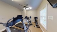 Located at 27642 US Highway 98 in Daphne, Alabama, the Arbors Fitness Center is a dedicated single-floor exercise facility designed to support an active lifestyle. The space is finished with neutral beige walls and low-pile carpeted flooring, creating a clean and functional environment conducive to focused workouts.

The fitness center is equipped with a comprehensive selection of cardio machines, including multiple True-brand treadmills, upright and recumbent exercise bikes, an elliptical cross-trainer, and a stair stepper. A wall-mounted flat-screen television is positioned for easy viewing during exercise, and a ceiling fan provides additional air circulation throughout the room.

Natural light filters in through several windows fitted with white blinds, lending the space a bright and welcoming atmosphere. Safety features include a fire extinguisher and illuminated exit signage near the keyed entry door, which leads directly to the exterior. Fitness center rules are posted prominently on the wall, and standard HVAC vents ensure comfortable climate control year-round.

The layout is thoughtfully organized to allow users to move between equipment with ease. Overall, the Arbors Fitness Center offers a practical, well-maintained amenity space suited for a range of cardiovascular exercise needs.