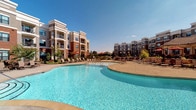 This outdoor amenity space is centered around a generously sized, freeform swimming pool with crystal-clear blue water, surrounded by a wide pool deck lined with lounge chairs for sunbathing and relaxation. The pool's curving design adds a resort-like quality to the environment, while the open sky above ensures an abundance of natural light throughout the day.

Adjacent to the pool, a covered pavilion provides a shaded retreat furnished with comfortable wicker seating, including sofas and armchairs with striped and solid cushions arranged around a decorative round table. This covered area features brick column accents, a smooth painted ceiling, and string lights that add ambiance during evening hours.

An open-air patio extends from the pavilion, offering additional seating with high-top tables and chairs shaded by bold red market umbrellas. The entire patio surface is finished in a refined multi-tone stone paver design that adds texture and visual interest underfoot.

Wrought iron fencing borders the space, providing a sense of enclosure while maintaining open sightlines to the surrounding landscaping and multi-story residential buildings. Greenery and flowering plants are thoughtfully integrated throughout, contributing to the overall inviting and well-maintained atmosphere of this outdoor gathering space.