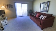 This single-story space features a living room designed for comfort and functionality. The room is finished with wall-to-wall carpeting in a neutral gray-blue tone, creating a soft and cohesive foundation. A large sliding glass door serves as a focal point, flooding the space with natural light and providing direct access to an outdoor balcony enclosed by a railing, where outdoor views extend beyond. Sheer vertical blinds frame the sliding door, allowing for adjustable light control while maintaining an airy feel. The living area offers ample open floor space, lending a sense of roominess suitable for various furniture arrangements. A wall-mounted mirror near the entry area adds visual depth to the room. The neutral-toned walls complement the overall understated palette of the space. The room's layout and generous natural light make it a welcoming and practical living environment, well-suited for everyday use and relaxation.