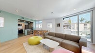 Located at 1503–1507 S Barrington Ave in Los Angeles, this single-floor common area offers a thoughtfully designed open-plan layout that seamlessly blends a living room and kitchen into one cohesive, communal environment. The living room is bright and airy, benefiting from large floor-to-ceiling windows and a sliding glass door that floods the space with natural light and provides views of the surrounding West LA neighborhood. Light hardwood flooring runs throughout, complementing the crisp white walls and clean-lined architecture. The living area is furnished with a generous sectional sofa, a modern low-profile coffee table, and playful donut-shaped accent seating in yellow and pink, adding a lively, social atmosphere. A flat-screen TV on a wooden media console and a pair of classic arcade game cabinets set against a sage-green accent wall add both entertainment and personality to the space. The kitchen features a contemporary design with light birchwood upper cabinetry, sage lower cabinets, white subway tile backsplash, and stainless steel appliances including a gas range, dishwasher, microwave, and refrigerator. A kitchen island with a light countertop provides additional prep and gathering space. A dining table with wooden chairs bridges the kitchen and living areas, completing this well-appointed, community-focused space.