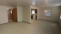 This single-floor home presents a straightforward and functional layout, with an open living and dining area that flows seamlessly into the kitchen. The living space is finished with wall-to-wall carpet in a neutral beige tone, providing a clean and understated foundation. An elegant chandelier hangs in the dining area near a window, adding a traditional touch to the space. The kitchen is accessible through a wide open passage and features medium-toned wood cabinetry, a white dishwasher, and a sink positioned beneath a window that allows natural light to filter in. The kitchen floor transitions to light-toned tile, offering a practical contrast to the carpeted living areas. Walls throughout are painted in a soft, neutral off-white, contributing to a bright and cohesive atmosphere. Dark wood baseboards frame each room, adding a subtle accent. A closet with warm wood-toned doors is visible along one wall, offering convenient storage. Floor vents are present throughout, indicating a forced-air heating and cooling system. Overall, the space is clean, practical, and well-suited for comfortable everyday living.
