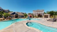 Located at 311 NW 96th St in Kansas City, Missouri, this single-level outdoor amenity space is designed to offer a resort-style experience within a vibrant residential community. The centerpiece is a generously sized swimming pool with crystal-clear blue water, complemented by an adjacent hot tub for year-round relaxation. The expansive concrete pool deck is lined with lounge chairs and shaded areas, providing ample space for sunbathing and socializing under open skies.

A covered outdoor patio serves as a comfortable gathering area, featuring a ceiling fan to keep the space cool during warm Missouri summers. The patio is furnished with a large sectional wicker sofa accented with colorful throw pillows, a coffee table, and additional seating, all arranged atop a decorative area rug. Large windows along the exterior wall allow natural light to flow into the adjacent interior space and offer views of the pool area.

The surrounding architecture blends stone accents, neutral lap siding, and pitched rooflines, creating a cohesive and attractive aesthetic throughout the community. Mature landscaping and greenery frame the space, adding a natural, welcoming ambiance. The overall design prioritizes outdoor living, leisure, and community engagement, making this amenity area a standout feature of the property.