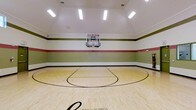 This single-level indoor basketball court offers a well-appointed space designed for serious recreational play. The court features a smooth, light-toned hardwood floor finished with regulation court markings, including a three-point arc and key, providing an authentic playing experience. A wall-mounted glass backboard with a standard hoop is positioned at one end of the court, built directly into the painted wall for a clean, integrated look.

The walls are decorated with horizontal color-block stripes in muted green, red, and white, lending a sporty yet polished aesthetic to the interior. Crown molding along the upper walls adds an unexpected touch of architectural refinement. The soaring ceiling height accommodates full gameplay comfortably, and the space is brightly illuminated by multiple recessed rectangular flush-mount light fixtures that ensure consistent, even lighting throughout the court. Ventilation is provided via ceiling vents, supporting comfort during active use. The overall design balances functionality with a well-finished interior, making this indoor court a standout amenity suited for year-round athletic activity.