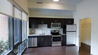 This single-floor space showcases a thoughtfully designed interior with a strong emphasis on modern aesthetics and functional living. The kitchen serves as a standout feature, boasting a full suite of stainless steel appliances including an electric range with oven, an over-the-range microwave, a dishwasher, and a full-size refrigerator. Rich, dark espresso-toned cabinetry lines the walls from floor to ceiling, providing ample storage while creating a bold visual contrast against the light-toned tile backsplash, which features a classic subway-style pattern. Granite or stone-look countertops add a refined, upscale touch to the workspace. The kitchen sink is positioned beneath upper cabinetry and is complemented by a sleek modern faucet. The flooring throughout is a deep, warm-toned hardwood, lending the space a grounded and cohesive feel. Large windows along one side of the unit allow natural light to filter in generously, enhancing the open and airy atmosphere. The elevated ceiling height adds a sense of volume and spaciousness to the overall layout. Together, these elements create a clean, contemporary living environment that balances style with practicality.