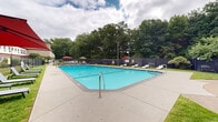 Located at 415 Porter Lake Dr in Springfield, Massachusetts, this outdoor recreational area centers around a generously sized swimming pool with clear turquoise water. The pool area is enclosed by a black chain-link fence and features clearly marked depth indicators, including a 6-foot depth designation, along with a no-diving policy. A concrete pool deck surrounds the water, providing ample space for movement and accessibility via stainless steel handrails.

Adjacent to the pool, a dedicated lounge area offers multiple reclining chaise lounges positioned on the surrounding lawn. A circular patio seating arrangement, shaded by large red cantilever umbrellas, provides a comfortable gathering spot for outdoor dining or relaxation. Additional white lounge chairs are arranged along the pool's perimeter for sunbathing.

The grounds are neatly landscaped with well-maintained grass and bordered by mature trees that provide a natural, green backdrop. Paved walkways connect the various areas of the space, leading to gated entry points with security signage. The multi-story brick building adjacent to the pool area features red accent trim and numerous windows overlooking the outdoor space, contributing to the overall functional and community-oriented character of this property.