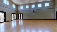 This community amenity space centers around a full indoor basketball court designed with both functionality and comfort in mind. The court itself is finished with polished hardwood flooring that reflects the abundant natural light streaming in from a row of clerestory windows positioned high along the upper walls. The soaring ceiling height creates an open, airy atmosphere well-suited for active play. A wall-mounted glass backboard and hoop are installed at regulation height, and black-trimmed double doors provide convenient access points on the court's perimeter.

Adjacent to the court building, a covered exterior entry area features rustic dark wood beam framing, stucco walls, and wall-mounted lighting, providing a transitional space between indoors and outdoors. Posted pool rules signal proximity to the community's aquatic amenities.

The outdoor pool area is a central gathering space, featuring a generously sized swimming pool alongside a separate spa. A broad concrete deck surrounds the water, furnished with lounge chairs and patio umbrellas. Mature trees offer natural shade, while views of the surrounding multi-story building create an enclosed, community-oriented atmosphere. Together, these amenities form a well-rounded recreational destination suited for both competitive activity and relaxed leisure.