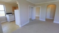 This single-floor space showcases a thoughtfully arranged layout that connects the main living area to adjacent rooms through wide, white-trimmed doorways and a decorative arched passage. Crown molding runs along the ceilings throughout, adding a classic architectural detail that elevates the overall aesthetic. The living area is finished with neutral-toned carpeting, contributing to a calm and cohesive atmosphere.

A half-wall partition separates the living space from the kitchen, which is visible through the arched opening and features warm wood-toned cabinetry, a white refrigerator, and a range. This semi-open design allows for visual connection between the spaces while maintaining some functional separation.

A mirrored sliding closet door provides storage within the living area while also reflecting light to enhance the sense of space. A separate doorway leads to an additional room, also carpeted and finished with matching white baseboards and trim, maintaining design consistency throughout. The neutral wall colors, white woodwork, and soft flooring create a clean, move-in-ready environment suitable for a variety of furniture arrangements and personal styles.