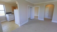 This single-floor space showcases a thoughtfully arranged layout that connects the main living area to adjacent rooms through wide, white-trimmed doorways and a decorative arched passage. Crown molding runs along the ceilings throughout, adding a classic architectural detail that elevates the overall aesthetic. The living area is finished with neutral-toned carpeting, contributing to a calm and cohesive atmosphere.

A half-wall partition separates the living space from the kitchen, which is visible through the arched opening and features warm wood-toned cabinetry, a white refrigerator, and a range. This semi-open design allows for visual connection between the spaces while maintaining some functional separation.

A mirrored sliding closet door provides storage within the living area while also reflecting light to enhance the sense of space. A separate doorway leads to an additional room, also carpeted and finished with matching white baseboards and trim, maintaining design consistency throughout. The neutral wall colors, white woodwork, and soft flooring create a clean, move-in-ready environment suitable for a variety of furniture arrangements and personal styles.