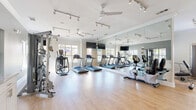 Located at 4045 George Busbee Pkwy NW in Kennesaw, Georgia, this fitness center offers a bright, well-appointed exercise space designed to support a comprehensive workout routine. The room features light hardwood-style flooring, crisp white crown molding, and a clean, modern aesthetic throughout.

The cardio area is lined with True-brand treadmills and elliptical trainers positioned in front of large windows that invite generous natural light and views of the surrounding neighborhood. Ceiling fans and track lighting overhead ensure the space remains comfortable and well-lit during any workout. Mounted flat-screen TVs provide entertainment options for cardio sessions.

A full wall of floor-to-ceiling mirrors visually expands the space while allowing users to monitor their form. The strength training section includes a multi-functional cable machine, a weight bench with free weights, and additional resistance equipment from brands such as Cybex, providing a thorough range of options for resistance training.

A rowing machine rounds out the cardio offerings, adding versatility to the equipment lineup. One wall features an eye-catching "live fit" graphic mural, reinforcing the motivational atmosphere of the space. A small service counter with cabinetry offers added convenience for users. The overall ambiance is bright, clean, and energizing — purpose-built to support an active lifestyle.