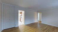 This single-floor residence offers a clean, well-maintained layout designed for comfortable everyday living. The main living area is characterized by warm, light-toned hardwood floors that run throughout, lending a sense of continuity and warmth to the space. Elegant crown molding lines the ceiling, adding a classic architectural detail that elevates the overall aesthetic. The neutral gray walls provide a versatile backdrop, allowing for a wide range of interior styling options.

A wide doorway opens directly into the kitchen, which features warm wood cabinetry, white appliances, and lighter flooring, creating a bright and functional cooking space. A second doorway leads to additional rooms beyond the main living area, suggesting a thoughtful and connected floor plan.

Natural light filters into the space, contributing to the airy and open atmosphere. The layout flows smoothly between rooms, making the residence feel cohesive and well-proportioned. Classic white baseboards and trim throughout complement the crown molding and reinforce the traditional detailing found across the home. Overall, the space combines timeless architectural character with practical functionality, making it a welcoming and adaptable living environment.