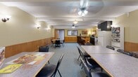 Located at 4440 Beard Ave S in Minneapolis, Minnesota, this community room serves as a versatile gathering and dining space. The single-floor layout offers a generous open area designed to accommodate a variety of social functions. Warm yellow walls, accented by wood wainscoting that lines the lower portion of every wall, lend the space a welcoming and classic character throughout.

The room is anchored by a built-in kitchenette featuring dark cabinetry, a dark granite-style countertop, a refrigerator, a microwave, and additional appliances — providing all the essentials for hosting events and shared meals. Adjacent to the kitchenette, a raised bar counter with dark wood paneling and leather-cushioned bar stools creates an inviting spot for casual gatherings.

The main floor area is carpeted in a soft blue-gray tone and arranged with multiple dining tables and chairs to seat a sizeable group. A separate lounge area with upholstered armchairs and side tables offers a relaxed setting for conversation. Ceiling fans with integrated light fixtures are distributed throughout the room, ensuring comfortable airflow and ambient lighting. Wall-mounted sconces add a warm, decorative glow. A window along one wall allows natural light to filter into the space, complementing the overall inviting atmosphere of this well-appointed community room.