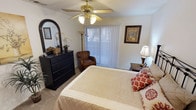 This single-floor space offers a warm and inviting atmosphere highlighted by thoughtful design and comfortable furnishings. The bedroom serves as a central feature, showcasing a ceiling fan with a brass finish and integrated lighting that adds both function and a classic aesthetic to the room. Vertical blinds adorn a large window, allowing natural light to filter in while providing privacy and light control.

The bedroom is furnished with a generously sized bed dressed in neutral, quilted bedding accented by decorative red and white patterned pillows, lending a cozy yet polished look. A dark wood chest of drawers paired with an arched mirror adds contrast and elegance, while a coordinating nightstand with a table lamp provides practical bedside lighting. A plush, carpeted floor adds warmth and comfort underfoot throughout the space.

A brown leather accent chair positioned near the window creates a relaxed reading nook, complemented by a tall floor lamp nearby. Framed wall art — including a classic map print — adds character and a timeless quality to the décor. The textured ceiling and clean white walls contribute to a bright and airy feel, completing this comfortable and well-designed living space.