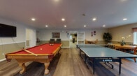 Located at 55 Golfview Dr in Manchester, NH, this single-floor community clubhouse offers a well-rounded recreational facility designed to serve residents with a variety of amenities. The space is divided into distinct areas, each catering to different interests and activities.

The fitness center is a standout feature, equipped with an extensive selection of cardio and strength-training equipment, including treadmills, elliptical machines, stationary bikes, and a multi-station cable machine. Wall-mounted mirrors and televisions enhance the workout experience, while large windows and recessed lighting keep the space bright and functional. Durable flooring throughout supports an active environment.

The game room provides an inviting space for recreation, featuring a full-size pool table with red felt, a ping-pong table, and comfortable seating areas. Wood-look flooring, recessed lighting, and decorative wall art contribute to a lively yet welcoming atmosphere. A dining set is also available nearby, adding versatility to the space.

The lounge area exudes warmth and comfort, anchored by a corner fireplace with a wood mantel and surrounded by plush leather seating. Crown molding, a ceiling fan, a large flat-screen television, and wood-look flooring complete the inviting setting, making it an ideal space for relaxation and socializing.