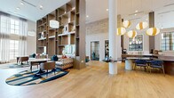 Located at 806 16th Ave N in Nashville's historic Marathon Village, The Scottie presents a generously proportioned single-floor communal space that blends modern design with warm, inviting character. The expansive open floor plan is anchored by wide-plank light oak hardwood flooring that flows seamlessly throughout, reflecting an abundance of natural light streaming in through oversized steel-framed windows that reach toward the dramatic double-height ceilings.

Exposed brick walls add a nod to the building's industrial heritage, contrasting beautifully with crisp white columns and contemporary finishes. A striking floor-to-ceiling built-in shelving unit serves as both a functional display feature and a visual focal point, while globe and orb pendant light fixtures add sculptural elegance overhead.

The lounge areas are thoughtfully arranged with eclectic, curated furnishings — including plush sofas, accent chairs, round tables, and colorful ottomans — creating distinct seating zones suited for socializing or relaxing. A central communal island with open shelving provides additional utility and display space. Abstract large-scale artwork adorns the soaring brick accent walls, lending a gallery-like quality. The grand double-door entry, framed by transom windows, makes for a memorable arrival experience throughout this vibrant Nashville destination.