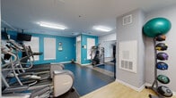 Located at 105 King Farm Blvd in Rockville, MD, this fitness center is thoughtfully divided into two connected workout areas, offering a well-rounded exercise environment. The space is finished with durable, multi-tone rubber flooring in the main workout zones and hardwood-style flooring in the entry and transitional areas, providing both function and visual variety.

The primary cardio area is energized by bold teal accent walls and is equipped with multiple treadmills, elliptical trainers, and a stair climber. Wall-mounted televisions are positioned for easy viewing during cardio sessions, and full-length mirrors along one wall enhance the sense of space. Flush-mount fluorescent lighting throughout ensures the entire facility is well-lit.

A secondary room houses a cable resistance machine, a rack of dumbbells, and a medicine ball storage rack, catering to strength and functional training needs. This area features neutral gray walls and large windows fitted with blinds, allowing natural light to filter in. A hand sanitizer station and posted instructional guides near the entrance reflect attention to hygiene and user guidance. Crown molding details add a refined touch to the overall aesthetic, making this fitness center a practical and inviting amenity for residents of the King Farm community.