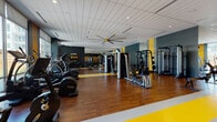 Located at 7545 E 29th Place in Denver's Central Park neighborhood, this fitness center offers a thoughtfully designed, single-floor space dedicated to comprehensive exercise and wellness. The interior features warm hardwood-style flooring accented by bold yellow and gray striped lane markings, which contrast against the dark charcoal and slate-toned walls to create a dynamic, energetic atmosphere. A drop-panel ceiling with recessed lighting and large ceiling fans ensures consistent airflow and even illumination throughout the space.

Floor-to-ceiling windows line one side of the facility, flooding the interior with natural light and providing open views of the surrounding neighborhood. The cardio zone is well-stocked with treadmills, elliptical trainers, stationary bikes, and stair climbers, all positioned to take advantage of the outdoor views. The strength training area includes cable machines, a multi-functional functional trainer frame, weight benches, a full dumbbell rack, and plate-loaded equipment. Full-length mirrors along one wall enhance the sense of depth and allow users to monitor their form during workouts. Medicine balls and accessory storage are neatly organized along the walls. The overall design is modern and professional, balancing functionality with an energizing aesthetic.