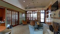 This single-floor lounge space offers a refined and comfortable environment designed for relaxed gathering and conversation. The room features elevated ceilings with recessed lighting and stylish pendant chandeliers that provide warm, layered illumination throughout. Exposed ductwork adds an industrial accent that contrasts tastefully with the space's otherwise polished aesthetic.

Large windows framed in rich wood trim line the exterior wall, fitted with horizontal blinds that allow for adjustable natural light and views of the outdoors. A glass-paneled entry door further enhances the connection to the exterior. The flooring is covered in a patterned carpet that adds texture and visual interest underfoot.

Seating is arranged in a conversational grouping, featuring a mix of teal upholstered armchairs and warm caramel-toned club chairs, creating an inviting and eclectic combination. Accent pillows add a decorative touch to the arrangement.

French doors with frosted glass panels on one wall provide access to an adjacent room while maintaining privacy. The walls are painted in soft sage green with a warm wood chair rail, contributing to an overall ambiance that feels both welcoming and professionally appointed.
