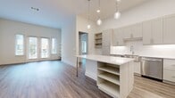 This well-appointed 2-bedroom, 2-bathroom single-story residence offers a thoughtfully designed open-concept layout finished with consistent wood-look flooring and a neutral color palette throughout. The kitchen serves as the heart of the home, featuring gray shaker-style cabinetry, a white quartz island with built-in shelving, a hexagonal tile backsplash, stainless steel appliances, and globe pendant lighting that adds a modern touch. The kitchen flows seamlessly into the living room, which is highlighted by soaring ceilings and a set of glass French doors that invite abundant natural light and outdoor views. Both bedrooms are generously sized with large windows fitted with blinds, tall ceilings, and easy access to closet storage, including a walk-in closet with dual hanging rods and shelving. The primary bathroom is a standout, offering a dual-sink vanity with gray cabinetry, a spacious glass-enclosed walk-in shower with bench seating, and large-format tile work. A dedicated laundry room provides full-size washer and dryer hookups with overhead shelving. Three hallways efficiently connect the living spaces, while two closets provide ample storage. The overall ambiance is clean, contemporary, and move-in ready.