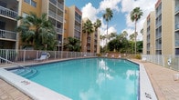 This outdoor communal space is defined by its expansive swimming pool with clear turquoise water, framed by a classic white metal fence and a wide brick-paved deck. The pool area is generously proportioned, offering ample room for lounging, with poolside chaise chairs positioned for relaxation along the perimeter.

The surrounding landscape is rich with mature tropical vegetation, including tall royal palms and fan palms that create a lush, resort-like atmosphere. Neatly mulched planting beds with ornamental shrubs line the fence interior, adding color and greenery throughout the space.

A dedicated seating and dining area adjacent to the pool features red metal picnic tables shaded by large blue market umbrellas, providing a comfortable outdoor gathering spot. The brick paver deck ties the entire space together with a warm, cohesive aesthetic.

The pool area is enclosed within a multi-story building complex finished in warm yellow and beige tones, with screened balconies overlooking the courtyard on all sides. The overall ambiance is tropical, bright, and welcoming, evoking the relaxed character of a South Florida setting. Natural light floods the space throughout the day, complemented by the open sky visible above the tree canopy.