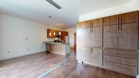This single-floor residential unit offers a well-designed, open-concept layout that seamlessly connects the kitchen and living areas. The kitchen is a standout feature, showcasing warm-toned wood cabinetry, granite countertops, and a full suite of stainless steel appliances including a refrigerator, electric range, and built-in microwave. A peninsula with pendant lighting overhead serves as both a prep space and casual dining area, complemented by track lighting above. Tall, floor-to-ceiling built-in cabinetry near the entry provides generous storage capacity. Rich hardwood-style flooring runs throughout the main living areas, adding warmth and continuity to the space. The living area benefits from a large picture window that fills the room with abundant natural light and offers views of the surrounding streetscape. A bathroom features tile flooring, a wood vanity with granite countertop, a large mirror, and modern bar-style hardware. The unit also includes an in-unit washer and dryer, adding everyday convenience. Clean white walls and a neutral palette throughout create a bright, functional environment suited to a range of uses.
