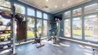 This single-floor fitness center is thoughtfully designed to support a comprehensive workout experience. The space is characterized by bold teal-blue walls accented with crisp white crown molding, creating a vibrant and energizing atmosphere. Expansive floor-to-ceiling windows line multiple walls, flooding the interior with natural light and offering open views of lush green landscaping and the surrounding community outdoors.

The cardio zone features Matrix-brand treadmills and an elliptical trainer positioned directly in front of the large windows, allowing users to enjoy scenic outdoor views during their workouts. A dedicated cycling station includes a connected smart spin bike for interactive training sessions. The strength training area is equipped with a multi-functional cable machine and an adjustable workout bench, ideal for a wide range of resistance exercises. Wall-mounted televisions are positioned throughout for entertainment.

A separate functional training section provides a polished concrete floor surface, a full-length mirror, a heavy punching bag for boxing and martial arts training, and a well-stocked rack of kettlebells, medicine balls, and dumbbells in various weights. Resistance bands and additional accessories are also available. The space is accessed through glass-paneled double doors and is finished with durable rubber flooring in the main exercise area, ensuring both safety and comfort during workouts.