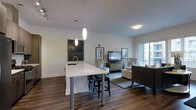 This single-floor residence offers a thoughtfully designed open-concept layout that seamlessly connects the kitchen and living areas. The kitchen is anchored by a striking white countertop island with a built-in sink, supported by polished steel legs, and illuminated by elegant pendant lights that hang from the smooth white ceiling. Bar-height stools line the island, making it an ideal spot for casual dining or entertaining.

Rich, dark hardwood flooring extends throughout the space, providing a warm contrast to the crisp white walls and bright surfaces. The living area is generously proportioned, featuring a comfortable seating arrangement centered around a media console. Framed artwork adorns the walls, adding a curated, contemporary feel to the room.

Natural light flows in through a large window and sliding glass door that opens onto a private balcony, offering views of the surrounding exterior. Recessed and flush-mount lighting complement the pendant fixtures, ensuring the space is well-lit throughout. The overall ambiance is clean, modern, and inviting, with a neutral color palette and thoughtful design details that create a cohesive and functional living environment.