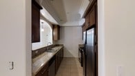 The kitchen serves as a focal point of this single-floor space, showcasing a galley-style layout that maximizes efficiency and functionality. Rich, dark wood cabinetry lines both sides of the kitchen, providing ample storage with brushed nickel hardware adding a refined touch. The countertops feature speckled granite in warm earth tones, offering both durability and visual appeal. Stainless steel appliances, including a French door refrigerator and a built-in range, contribute a modern, professional aesthetic.

A double-basin stainless steel sink is positioned along the left counter run beneath recessed lighting, while pendant lights visible through an arched pass-through illuminate an adjacent area, suggesting an open connection to a neighboring room. The flooring consists of neutral-toned ceramic tiles that complement the overall warm color palette.

A notable architectural detail is the tray ceiling with crown molding, which adds dimension and elegance to the kitchen space. The arched opening adds a classic architectural element, seamlessly transitioning between spaces while maintaining a cohesive design throughout.