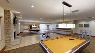 This single-floor recreational space is designed to provide a variety of leisure and social activities within a modern, open-plan layout. The room is generously sized, with recessed lighting throughout the ceiling supplemented by pendant fixtures hanging above the pool tables, casting focused light over the playing surfaces. Two full-sized pool tables are positioned prominently within the space, with a ping-pong table centrally located to encourage active play. The flooring transitions between tile and patterned carpet, helping to visually define different zones within the room. A curved reception or service counter is situated toward one side, featuring a two-tone design that adds a contemporary touch. In the background, a secondary area is visible, furnished with bar-height seating and additional recreational equipment, suggesting a multi-zone layout ideal for group gatherings. The neutral color palette of whites, beiges, and grays gives the space a clean, welcoming atmosphere. The overall design is functional yet comfortable, making it well-suited as a shared amenity area for socializing and entertainment.