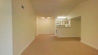 This single-floor residence offers a straightforward and functional layout designed for comfortable everyday living. The main living area is generously proportioned, featuring wall-to-wall carpet flooring in a neutral beige tone that lends warmth to the space. Smooth, cream-colored walls and white baseboards keep the aesthetic clean and understated throughout.

A key feature of the layout is the open connection between the living area and the kitchen, separated by a pass-through counter that allows for easy interaction between the two spaces. The kitchen is equipped with white cabinetry accented by wood-toned trim, providing a classic contrast. A fluorescent ceiling fixture provides direct illumination over the kitchen workspace.

The living area benefits from recessed-style track lighting mounted on the ceiling, ensuring even distribution of light. Vents are visible along the upper wall and ceiling, indicating a centralized HVAC system for climate control. A doorway leads to additional rooms, suggesting a private sleeping or utility area tucked away from the main living space. The overall ambiance is calm and neutral, offering a blank canvas well-suited for personalization.