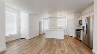 This two-story home presents a bright, contemporary design characterized by a crisp white palette, light wood-style luxury vinyl plank flooring, and an open, airy layout across both levels. The main floor centers on an open-concept living and kitchen area, where the two spaces flow seamlessly into one another. The kitchen is well-appointed with white shaker-style cabinetry, light countertops, a stainless steel sink, and a full suite of stainless steel appliances including a gas range, over-the-range microwave, and refrigerator. A central island provides additional prep and seating space, while pendant lighting with brushed gold accents adds a modern touch. Natural light fills the main floor through multiple windows fitted with horizontal blinds. A staircase leads to the upper level, where carpeted bedrooms provide comfort underfoot. The primary bedroom features a walk-in closet with built-in shelving and rod systems, offering generous storage. The bathroom includes a full tub-shower combination with clean white surround panels and wood-look flooring. Throughout the home, white shaker-style interior doors, clean baseboards, and a neutral color scheme create a cohesive, move-in-ready aesthetic suited for a variety of lifestyles.