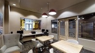 This single-floor workspace strikes a balance between functionality and contemporary design, making it well-suited for collaborative and professional environments alike. The open seating area is furnished with light natural wood tables and modern upholstered chairs, creating an inviting atmosphere for meetings, focused work, or casual gatherings. Overhead, bold red pendant lights with a vintage-industrial aesthetic serve as a striking focal point, casting warm, even illumination throughout the space.

The interior features a mix of glass partitions and sliding wood-framed doors, allowing natural light to flow through while maintaining defined zones for privacy and concentration. A built-in workstation area lines one wall, equipped with monitors and workspace essentials, offering a practical solution for shared office use.

The flooring features patterned carpet tiles in dark, muted tones that complement the warm wood tones of the furniture and doors. Frosted glass panels bearing a distinctive logo add a branded, professional touch to the space. The overall ambiance is polished yet approachable, combining industrial-inspired accents with warm natural materials to create a workspace that is both productive and visually engaging.