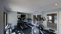 This single-floor fitness space is designed to provide a practical and focused exercise environment. The room is outfitted with multiple treadmills and elliptical machines, offering a solid selection of cardio equipment suited for a range of fitness routines. The dark rubberized flooring provides both durability and stability underfoot, a practical choice for a high-use exercise space.

A wall of mirrors along one side visually expands the room while allowing users to monitor their form during workouts. A flat-screen television is mounted on the mirrored wall, adding an entertainment element to cardio sessions. Recessed lighting in the smooth ceiling provides consistent, even illumination throughout the space.

Natural light filters in through multiple windows fitted with horizontal blinds, contributing to a bright and open atmosphere without compromising privacy. The walls are painted in a neutral gray tone, giving the room a clean and modern aesthetic. The overall layout is efficient, making good use of the available space to accommodate equipment while maintaining comfortable circulation between machines.