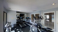 This single-floor fitness space is designed to provide a practical and focused exercise environment. The room is outfitted with multiple treadmills and elliptical machines, offering a solid selection of cardio equipment suited for a range of fitness routines. The dark rubberized flooring provides both durability and stability underfoot, a practical choice for a high-use exercise space.

A wall of mirrors along one side visually expands the room while allowing users to monitor their form during workouts. A flat-screen television is mounted on the mirrored wall, adding an entertainment element to cardio sessions. Recessed lighting in the smooth ceiling provides consistent, even illumination throughout the space.

Natural light filters in through multiple windows fitted with horizontal blinds, contributing to a bright and open atmosphere without compromising privacy. The walls are painted in a neutral gray tone, giving the room a clean and modern aesthetic. The overall layout is efficient, making good use of the available space to accommodate equipment while maintaining comfortable circulation between machines.