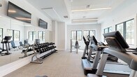 This fitness center offers a comprehensively equipped exercise space designed with both functionality and aesthetics in mind. The room features a clean, modern aesthetic with crisp white walls, a coffered ceiling with integrated linear lighting, and neutral-toned carpet tiles that provide a practical and durable workout surface.

Natural light flows generously through large, black-framed windows positioned along multiple walls, offering pleasant outdoor views of surrounding greenery and keeping the space feeling open and airy. Full-length mirrors mounted on one wall visually expand the room while allowing users to monitor their form during exercise.

The cardio zone is outfitted with a row of Life Fitness treadmills and elliptical machines, each equipped with individual display screens. Adjacent to these, a dedicated strength training area houses multiple selectorized cable and resistance machines, including a leg press and cable station, catering to a full range of muscle groups. A multi-tiered dumbbell rack stocked with a complete set of free weights rounds out the strength offerings. A power rack with a flat bench provides additional options for compound lifts.

Wall-mounted flat-screen televisions are positioned at strategic locations throughout the room, ensuring visibility from both the cardio and strength zones. The overall design is bright, uncluttered, and purpose-built for an effective workout experience.