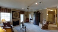 This single-floor residence exudes a classic, traditional charm that is both welcoming and refined. The main living area serves as the heart of the home, featuring a tile-surround fireplace that anchors the room with warmth and character. Plush wall-to-wall carpeting runs underfoot, complementing the warm, neutral-toned walls that create an inviting ambiance throughout.

Crown molding frames the ceilings with an elegant finishing touch, while recessed lighting and a classic ceiling fan with light kit provide both functional illumination and a sense of style. A glass-top accent table with ornate metalwork legs adds a decorative focal point to the seating area, which is appointed with traditional upholstered furniture.

A large, dark wood armoire serves as a striking storage piece, contributing to the room's rich, classic aesthetic. Decorative wall art and candle accents further enhance the traditional character of the space. A doorway visible from the living area leads to what appears to be a dedicated workspace or study, offering convenient access to additional functional areas of the home.

Natural light filters in through glass-panel doors, brightening the space and providing a connection to the outdoors. Overall, this home presents a cohesive, traditionally styled interior that balances comfort and elegance.