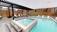 This single-story amenity space is designed to offer residents a well-rounded indoor-outdoor experience. The outdoor area features a rectangular swimming pool alongside an elevated hot tub finished in neutral tile, bordered by a smooth pool deck outfitted with white lounge chairs. A sleek black pergola provides shaded seating above the hot tub, while horizontal wood privacy fencing encloses the space, creating a sense of seclusion. An adjacent patio area includes dining furniture and greenery, adding a landscaped element to the outdoor environment.

The interior lounge is bright and open, featuring large-format tile flooring, recessed lighting, and ceiling fans that maintain air circulation throughout. Bold orange upholstered armchairs with accent pillows are arranged around white coffee tables, creating a casual and inviting seating area. A small beverage station is positioned along one wall, enhancing the functionality of the space.

Floor-to-ceiling glass partition walls and black-framed sliding glass doors visually and physically connect the interior lounge to both an adjoining room and the outdoor patio areas. The extensive use of glass promotes an abundance of natural light and maintains sightlines throughout the space, contributing to a cohesive and airy atmosphere overall.