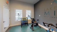 This dedicated fitness studio, managed by Greystar, is purpose-built to support a well-rounded workout experience within a single, thoughtfully designed space. The room features a teal-toned interlocking floor mat surface that defines the workout area, providing cushioning and support for high-intensity exercise. A fleet of Stages indoor cycling bikes is arranged across the floor, making the space ideal for spin classes or solo cycling sessions. A large wall-mounted display screen faces the riders, enabling guided virtual workouts and immersive cycling content. Natural light filters in through multiple windows, offering views of lush greenery and landscaping outside, contributing to an energizing atmosphere. Recessed ceiling lighting supplements the natural light, ensuring the space remains bright and functional at any hour. A wooden rolling utility rack along one wall provides convenient storage for rolled towels and accessories. A free weights station features a tiered dumbbell rack stocked with color-coded weights, accompanied by an additional spin bike for individual use. Step platforms and stretching equipment round out the available gear. The studio connects directly to an adjacent fitness area visible through interior windows, extending the overall workout facility. The two-tone gray and white wall design adds a modern, motivating aesthetic throughout.