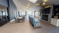 This single-floor community clubhouse offers a generously proportioned, open-plan interior designed for social gathering and recreation. The main hall features soaring vaulted ceilings with a skylight, track lighting, and large windows lining the far wall that invite abundant natural light and outdoor views. The flooring throughout is a light wood-tone laminate that enhances the airy, welcoming atmosphere.

A central lounge area is anchored by a standout fireplace wall clad in decorative patterned tile, framing a large mounted television and flanked by gold accent décor. Plush blue upholstered seating faces the fireplace, creating a comfortable gathering focal point. Adjacent to the lounge, a dining area provides multiple wood-and-upholstered chair sets arranged around rectangular tables suited for group meals or activities.

A ping-pong table is positioned near the windowed wall, adding a recreational element to the space. Dark structural columns define zones within the open layout without disrupting visual flow. A compact, clean-lined kitchenette with white cabinetry and a prep counter supports event hosting. Dark-framed glass sliding doors partition a separate flex room, while a welcoming entry foyer with framed artwork and a console table completes the space.