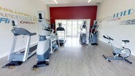This fitness center offers a dedicated space for cardio training and exercise, thoughtfully designed to motivate and energize its users. The room features light wood-tone flooring that creates a clean, modern aesthetic, while a bold crimson accent wall at the far end serves as a striking visual focal point. Natural light pours in through a set of glass-paned French doors centered on that wall, offering views of the outdoors and contributing to a bright, welcoming atmosphere. The space is outfitted with a variety of cardio equipment, including elliptical machines, treadmills, and step trainers, arranged to allow comfortable circulation throughout the room. Motivational typography and fitness-themed graphics adorn the surrounding walls, reinforcing an active and goal-oriented ambiance. Recessed and linear lighting fixtures in the ceiling ensure consistent illumination throughout the space. The overall design balances functionality with an uplifting environment, making it an ideal setting for focused workouts. The single-floor layout ensures easy accessibility, and the open floor plan allows for an unobstructed, spacious feel that accommodates multiple users simultaneously.
