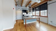 This thoughtfully designed industrial-modern loft seamlessly merges raw architectural character with contemporary finishes. Exposed timber beams traverse the ceiling throughout, complemented by visible ductwork and white shiplap-style ceiling panels that together create a distinctive warehouse-inspired aesthetic.

The open main living area is anchored by wide-plank hardwood floors that run continuously through the space, reflecting abundant natural light that pours in through a generous bank of multi-pane windows framed in warm wood trim. A bold blue accent wall adds a striking focal point to the living area, while industrial-style pendant lighting hangs from the beamed ceiling above.

The kitchen is a standout feature, equipped with dark cabinetry, stainless steel appliances including a refrigerator and dishwasher, a farmhouse-style stainless sink, open stainless shelving, and a classic white subway tile backsplash. A large central island with a concrete-style countertop doubles as a dining table extension, making it ideal for entertaining.

Additional rooms include flexible open spaces suitable for sleeping or office use, each continuing the hardwood flooring and exposed-beam ceiling details. Double Shaker-style closet doors provide ample storage. The bathroom offers a dual-sink vanity with a wood-framed mirror, light tile flooring, and a glass-enclosed walk-in shower with white subway tile and a rainfall showerhead, completing this refined yet industrial retreat.