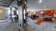This single-floor fitness center offers a well-appointed exercise space outfitted with professional-grade equipment and a striking visual identity defined by bold orange and grey tones. The flooring features patterned carpet tiles with vibrant orange accents that echo throughout the room, creating a cohesive and energizing aesthetic. Recessed ceiling lighting provides consistent, even illumination across the entire space.

The facility is equipped with a Synergy360 functional training system, a prominent centerpiece that supports a wide variety of cable and resistance-based exercises. Complementing this is a range of strength machines finished in matching orange and black, including what appears to be a leg press and multiple selectorized units. Cardio equipment, including treadmills, is positioned toward the rear of the space.

The layout is thoughtfully organized, with dedicated zones for strength training and open floor areas that allow for functional movement and stretching. An accent wall featuring bold graphic artwork adds visual depth and reinforces the dynamic character of the space. The overall ambiance is modern, motivating, and purpose-built for fitness.